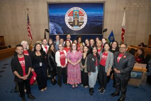 A large group of people, some wearing red shirts with "RTI" printed on them, gather in a government chamber in front of the official Los Angeles County seal for a group photo.