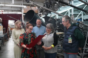 Five individuals stand together inside a warehouse filled with mechanical equipment, as one person reads from a red certificate or proclamation while the others look on attentively.