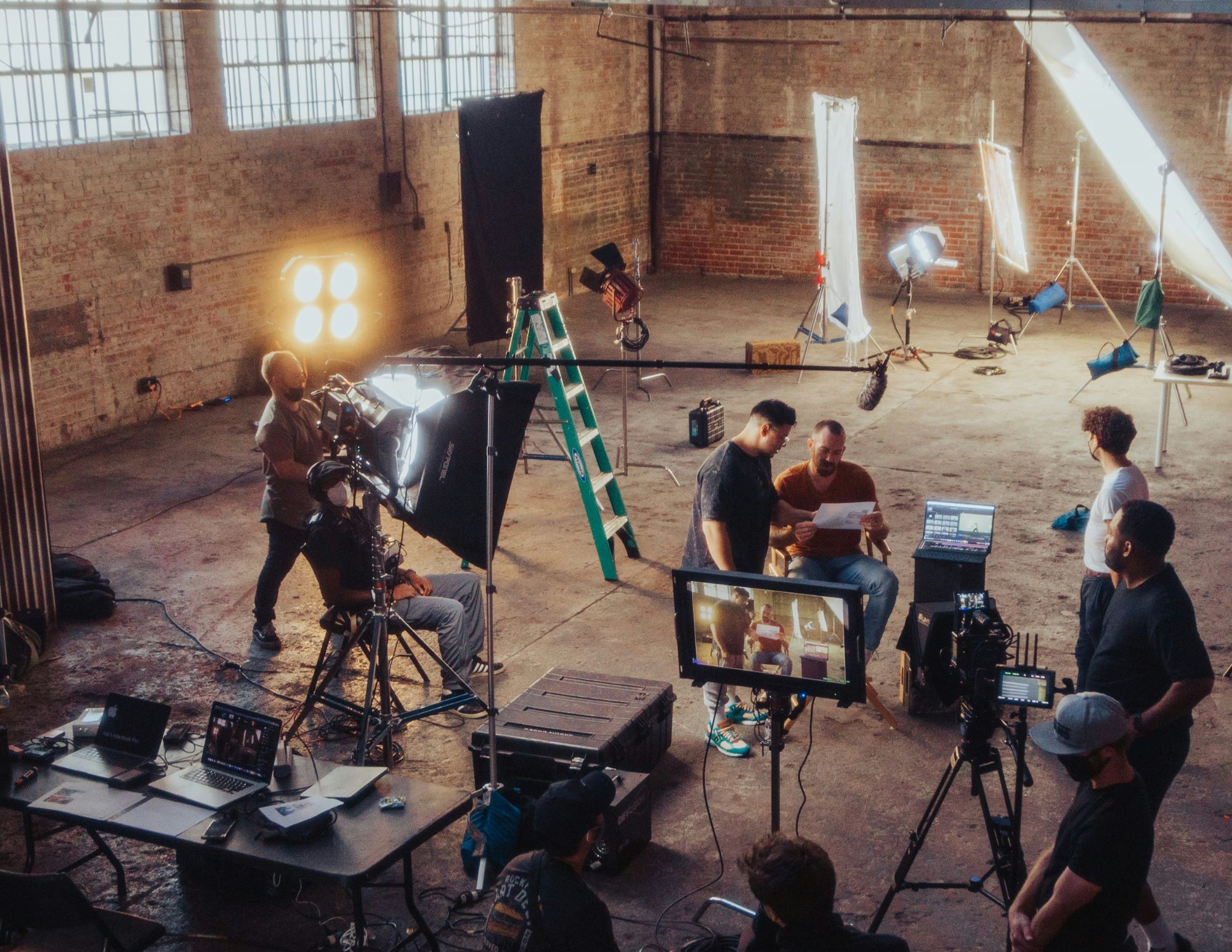 Behind-the-scenes view of a film or video production set in a large industrial studio with professional lighting, cameras, crew members, monitors, and equipment preparing for a shoot.