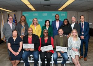 A group photo of fifteen individuals posing indoors, with three people in the front row holding large ceremonial checks. The group stands in front of a teal backdrop with Los Angeles County Department of Economic Opportunity branding.