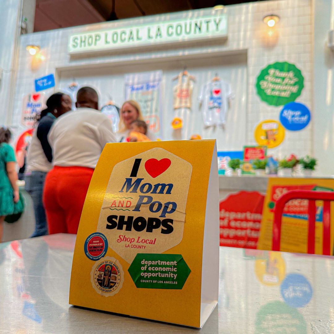 Table sign reading “I love Mom and Pop Shops – Shop Local LA County” is displayed in front of a colorful vendor booth promoting local businesses, with people interacting in the background.