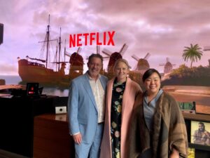Three people pose for a photo in front of a large screen displaying the Netflix logo with a scenic background of ships, windmills, and palm trees.
