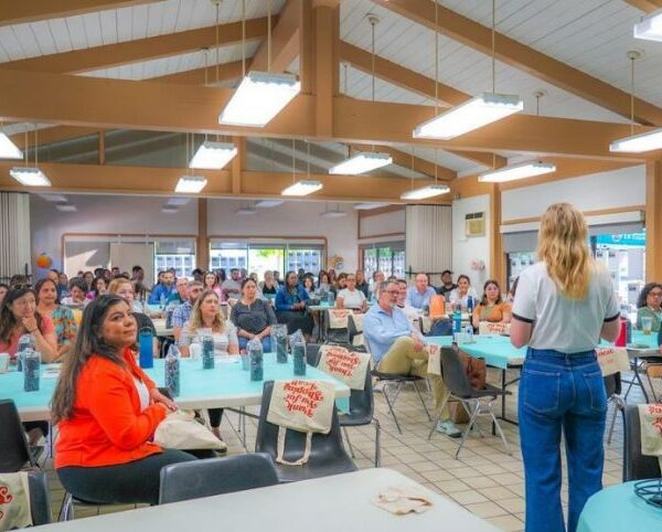 A woman stands addressing a full room of seated attendees during a community meeting or workshop, held in a bright hall with exposed wooden beams and round tables covered in teal tablecloths and giveaway bags.