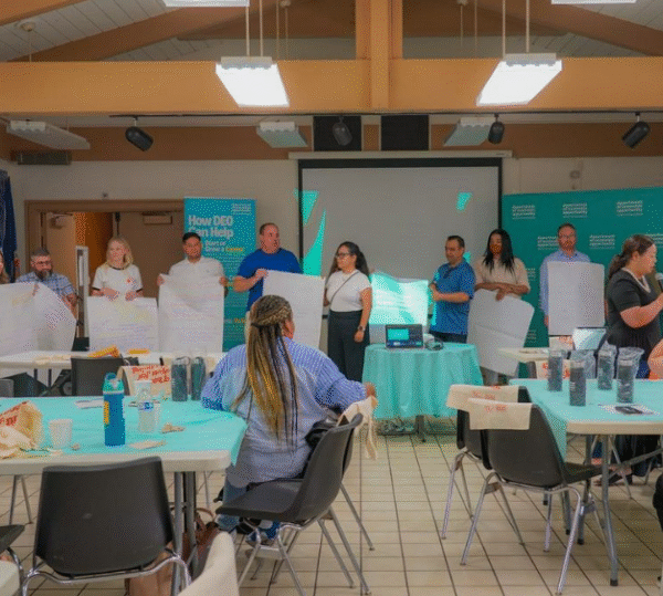 Group of people standing at the front of a community meeting room holding large sheets of paper while presenting ideas, with attendees seated at tables labeled with Department of Economic Opportunity materials.