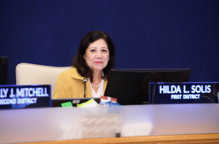 Supervisor Hilda L. Solis seated at a government meeting desk with a nameplate that reads “Hilda L. Solis, First District,” smiling and wearing a yellow blazer.