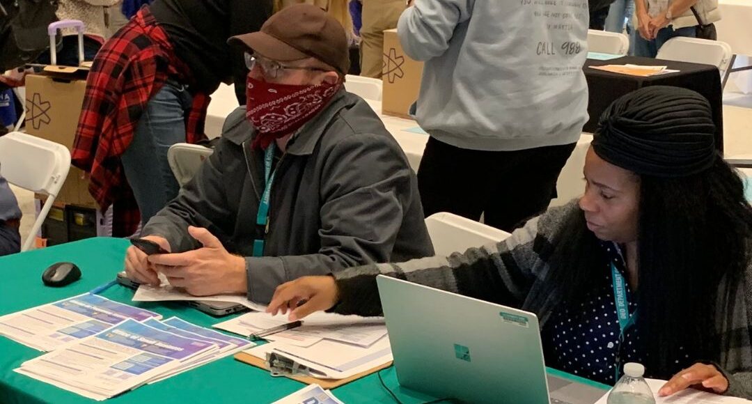 Two individuals sit at an assistance table during a community event, reviewing documents and using a laptop, with printed brochures and a green tablecloth in front of them.