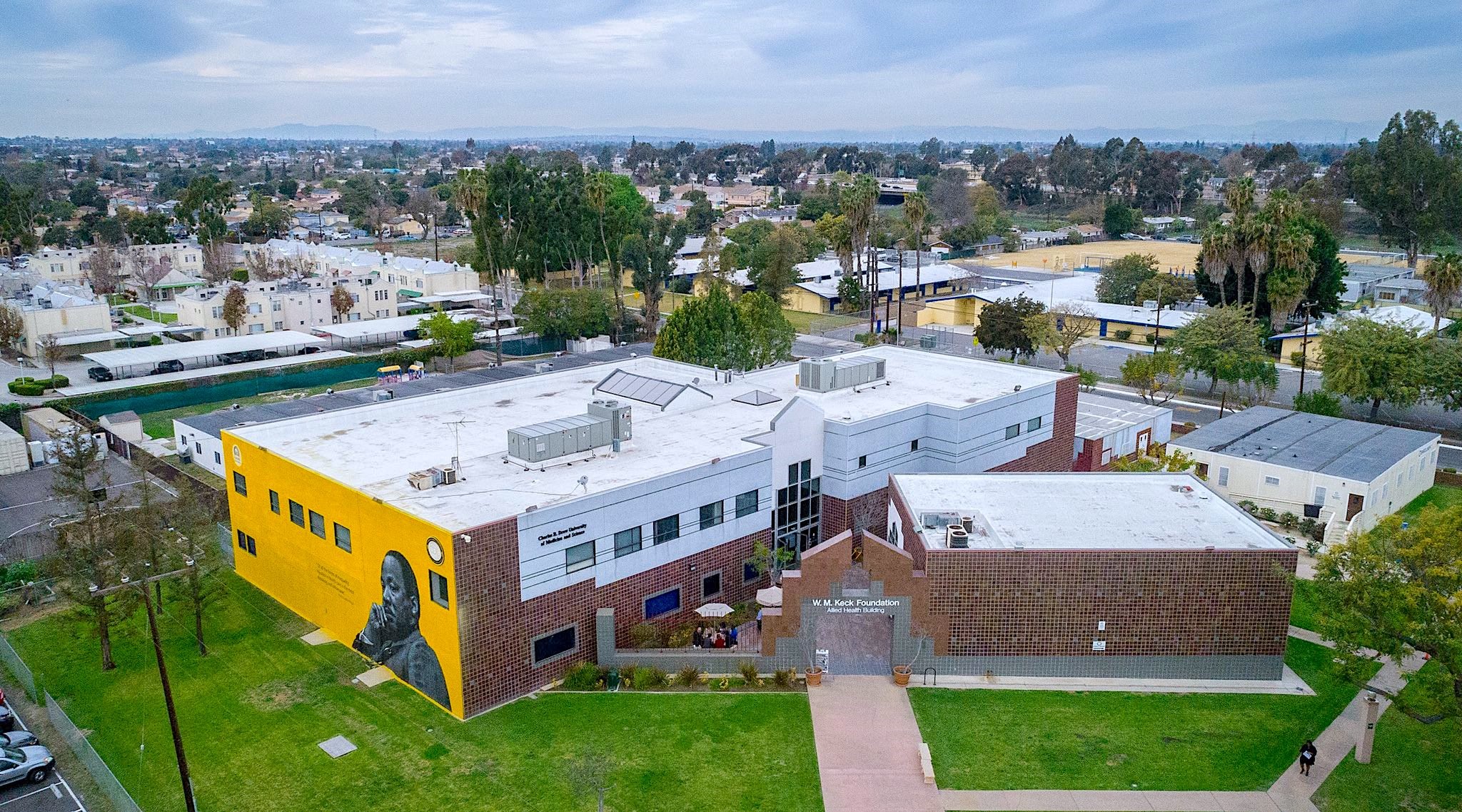 Aerial view of the W.M. Keck Foundation Allied Health Building at Charles R. Drew University, featuring a yellow mural wall with a portrait, surrounded by residential buildings, trees, and an open field under a cloudy sky.
