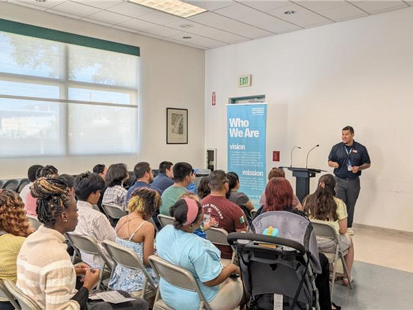 A group of attendees seated in rows at a community job center listening to a presenter standing at the front of the room near banners that read 'Who We Are' and 'America’s Job Center of California.