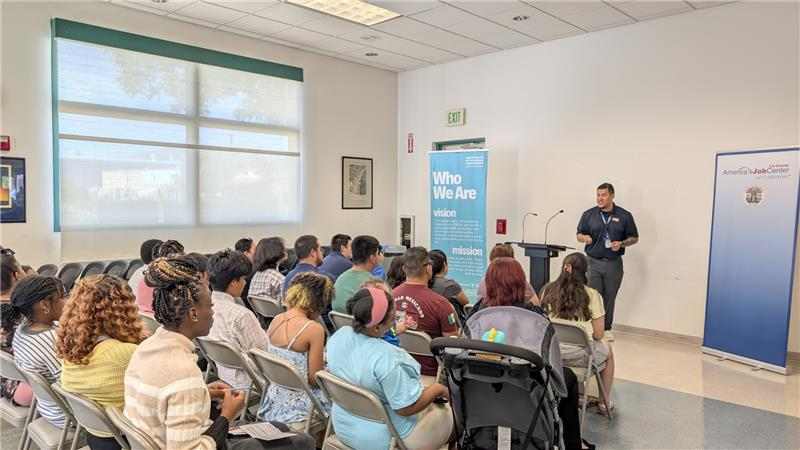 A group of attendees seated in rows at a community job center listening to a presenter standing at the front of the room near banners that read 'Who We Are' and 'America&rsquo;s Job Center of California.