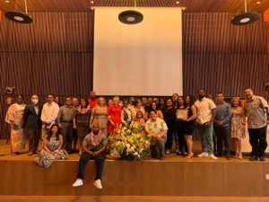 A large group of people poses together on stage in front of a flower arrangement and a large projector screen, suggesting a celebratory or graduation event.