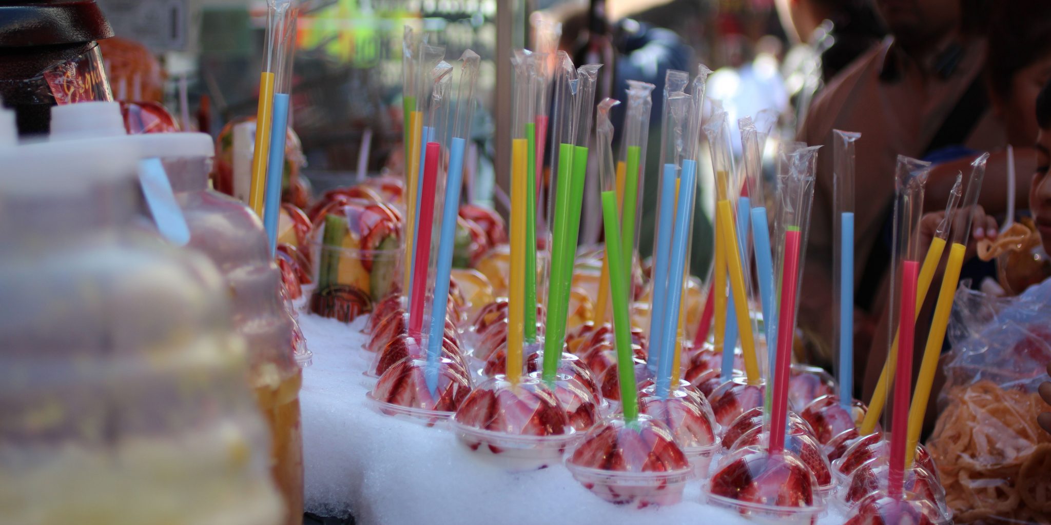 Clear plastic cups filled with sliced strawberries are arranged on a vendor&rsquo;s table, each covered with a clear lid and containing a colorful straw in sealed plastic wrapping.