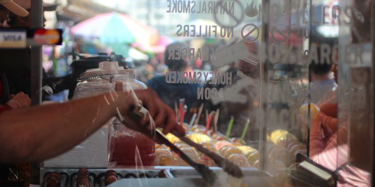 A vendor uses tongs to prepare food behind a glass panel with etched text, with colorful drinks and fruit cups visible in the background at an outdoor market.