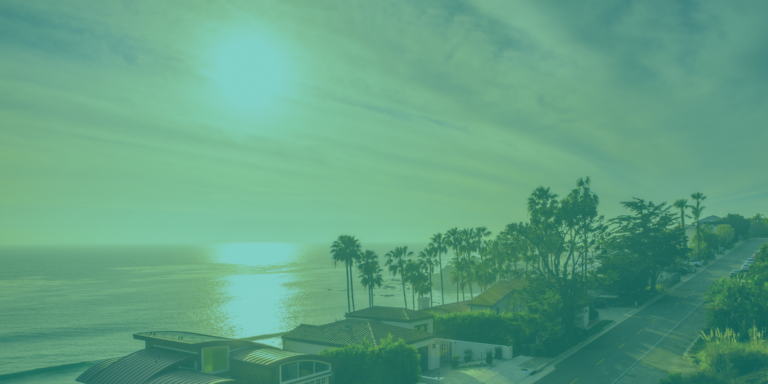 A coastal road lined with palm trees and homes overlooks the ocean at sunset under a greenish-blue tint.