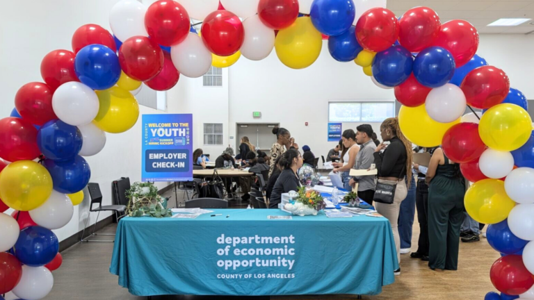 A colorful balloon arch in red, white, blue, and yellow frames the entrance to a Youth@Work event hosted by the Department of Economic Opportunity, County of Los Angeles. A check-in table with a teal tablecloth and program staff sits at the front, while attendees engage with employers in the background. A sign reads "Welcome to the Youth@Work Employer Check-In.