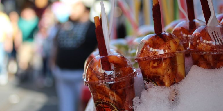 Cups filled with sliced fruit coated in chili powder, topped with tamarind sticks, and served with plastic forks, displayed on ice at an outdoor market.