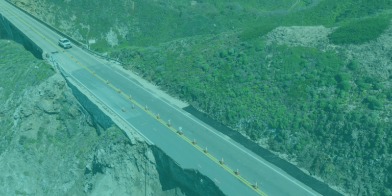 Aerial view of a damaged mountain road where a large section has collapsed, leaving a deep gap. A white vehicle is seen stopped near the edge, and traffic cones line the remaining portion of the road to block access. The surrounding area is covered in green vegetation and steep rocky terrain.