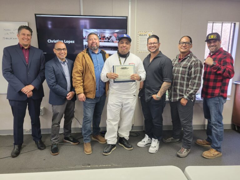 A group of men stands together indoors, with the person in the middle holding a certificate while smiling, as others beside him also smile and pose proudly for the photo.