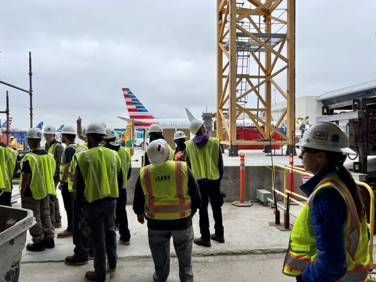 Workers in safety vests and hard hats gather at a construction site near an airport runway, with visible airplanes in the background.