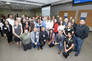 A large group of people is gathered together indoors for a group photo at an event, with individuals smiling and dressed in both professional and casual attire, some wearing name badges, standing in front of presentation screens and display boards in a conference room setting.
