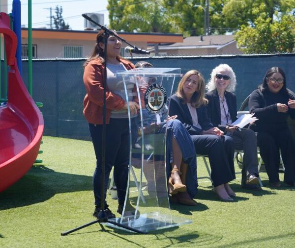 A woman speaks at a podium in front of a playground as four seated women listen and smile during an outdoor event.