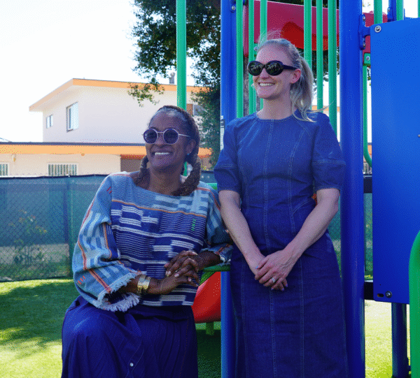 Two women smile while standing in front of a colorful playground structure on a sunny day, with one leaning casually against a play feature.