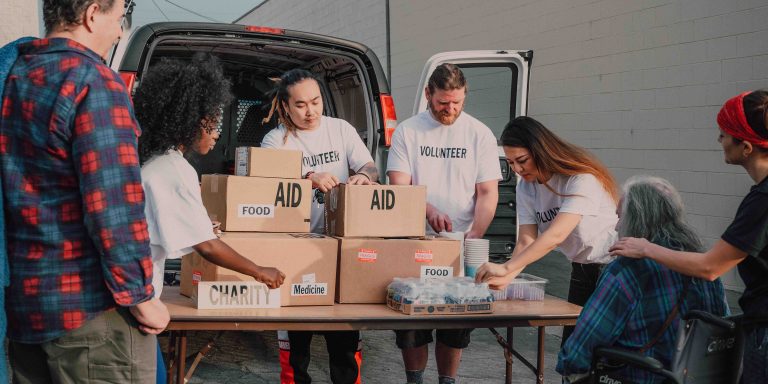 A group of volunteers wearing white "Volunteer" shirts unloads boxes labeled “AID,” “FOOD,” “CHARITY,” and “Medicine” from a van and organizes supplies on a table while distributing aid to community members, including an elderly person in a wheelchair.