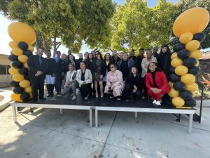 A group of people pose for a photo on an outdoor stage decorated with black and gold balloon columns, surrounded by trees and sunlight.