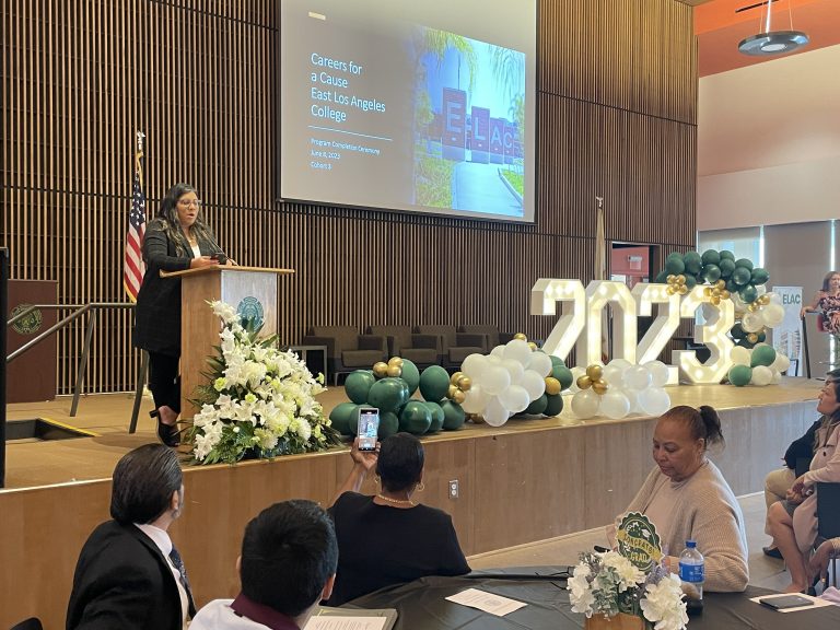 A woman speaks at a podium decorated with white flowers during a graduation event at East Los Angeles College, with a presentation slide and large &ldquo;2023&rdquo; balloon display in the background as attendees watch and take photos.