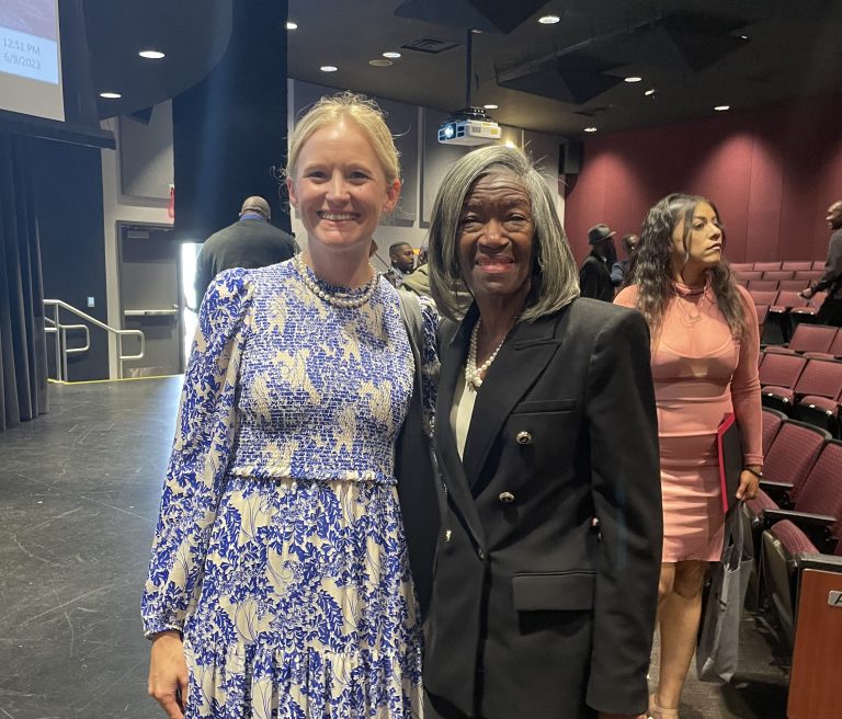 Two women smile for a photo inside an auditorium, one wearing a blue and white floral dress and the other in a black suit, with people and theater seating visible in the background.