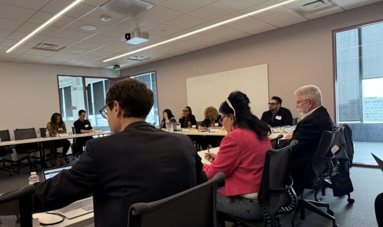 A group of professionals sit around a large conference table in a modern meeting room, actively engaged in discussion, taking notes, and working on laptops, with nameplates, notebooks, and water bottles in front of them.
