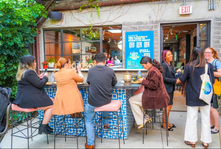 People sit and stand around an outdoor bar with blue patterned tiles, enjoying food and conversation, while a large informational sign for the East LA Entrepreneur Center is displayed on the wall in the background.