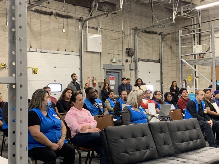 A group of people, many wearing blue vests, sit attentively in a warehouse setting during a presentation, with some taking notes and others observing from the back of the room.