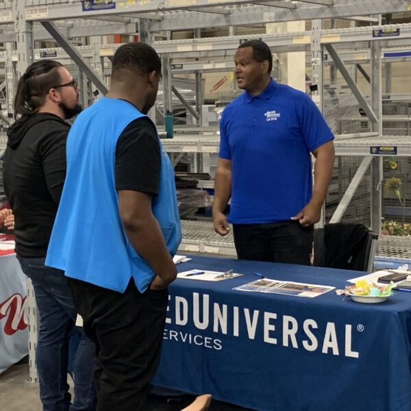 Two men speak with a recruiter at an Allied Universal booth during an event held in a warehouse setting.