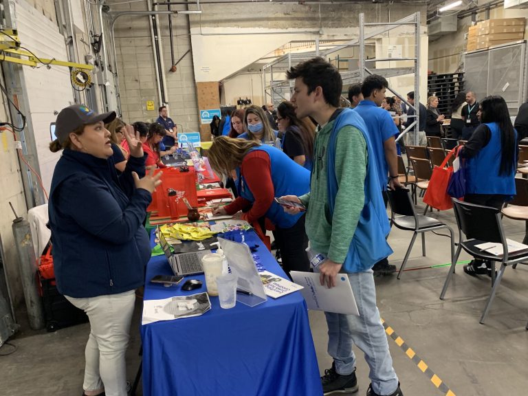 A woman speaks with a young man holding papers at a booth during a job fair in a warehouse setting, while other participants in blue vests engage with different tables in the background.