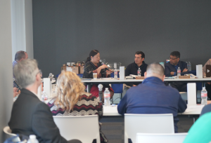 People seated around conference tables engage in a group discussion, with one woman speaking while others listen attentively in a professional meeting setting.