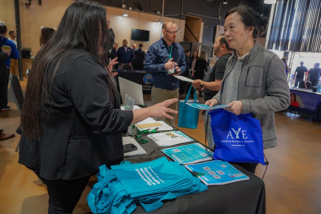 A woman speaks with an attendee holding a blue AYE tote bag at an indoor event booth covered with Department of Economic Opportunity materials and teal giveaway bags.