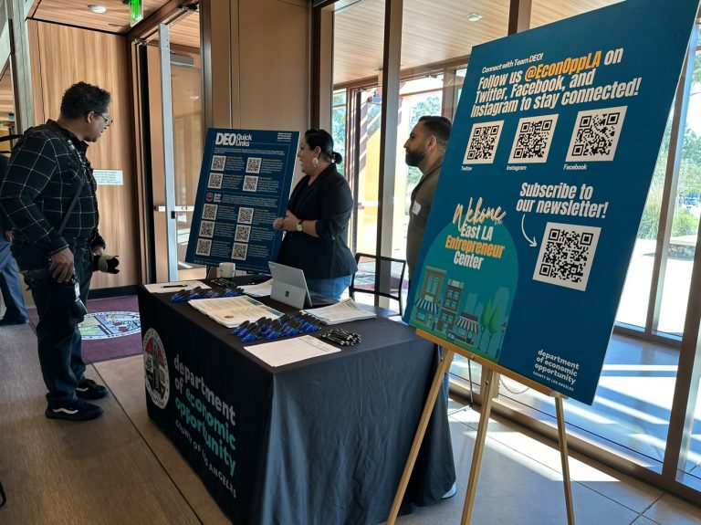 A man speaks with two representatives at a Department of Economic Opportunity booth featuring large boards with QR codes for resources, social media, and newsletter sign-ups inside a bright, wood-paneled venue.