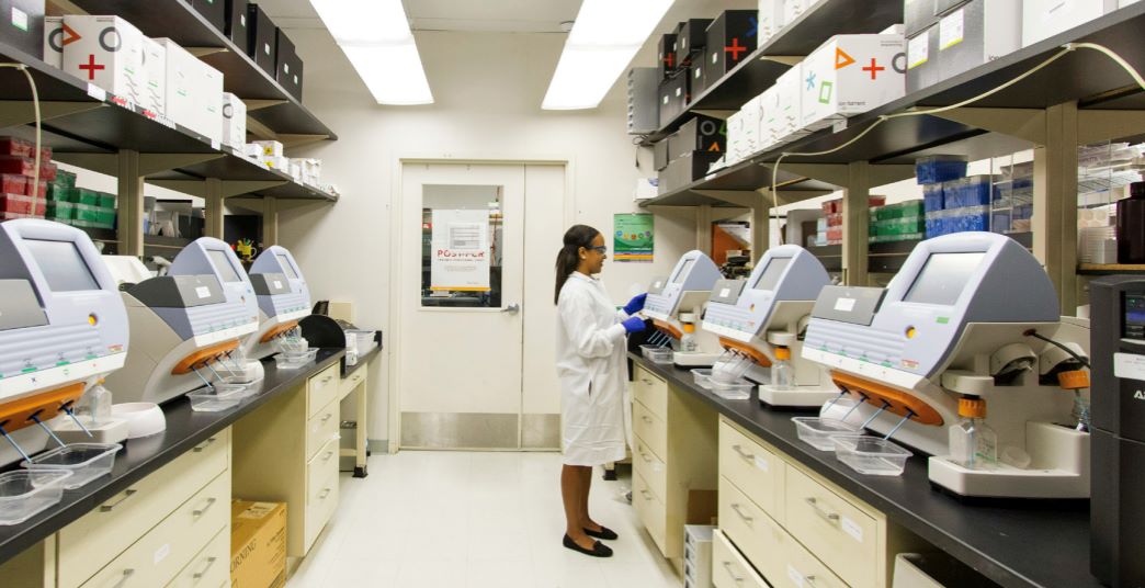 A scientist in a white lab coat and gloves works in a narrow laboratory room lined with automated machines on both sides. Shelves above the counters hold organized lab supplies and equipment, and a door at the end of the room leads to another area.