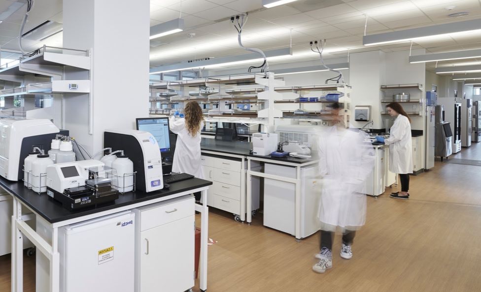 Modern laboratory with multiple researchers in white lab coats working at stations equipped with scientific instruments, computers, and lab equipment. The space is clean, bright, and organized, with shelves holding supplies and a wood-toned floor. One person is blurred while walking, indicating motion.