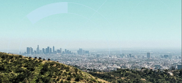 A wide view of the Los Angeles skyline on a clear day, seen from a hillside with greenery in the foreground and high-rise buildings stretching across the horizon.