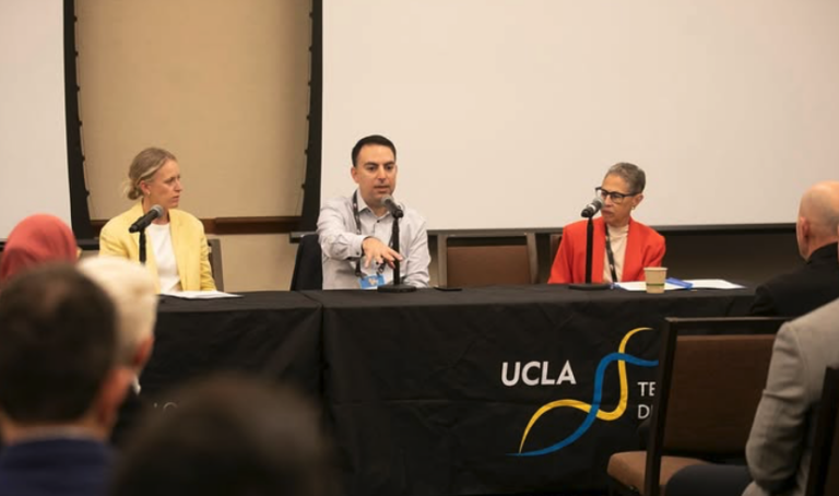 Panel discussion with three speakers seated at a table covered with a UCLA banner. The atmosphere is professional, with attentive audience members.