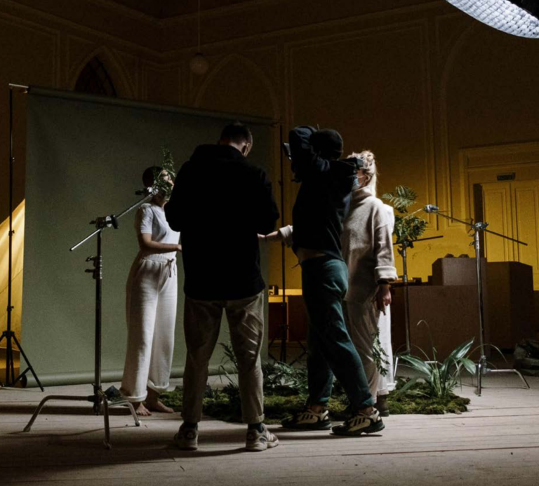 A group of people is on a dimly lit set with lighting equipment, microphones, and plants arranged on the floor, as they prepare or rehearse a scene in front of a backdrop.