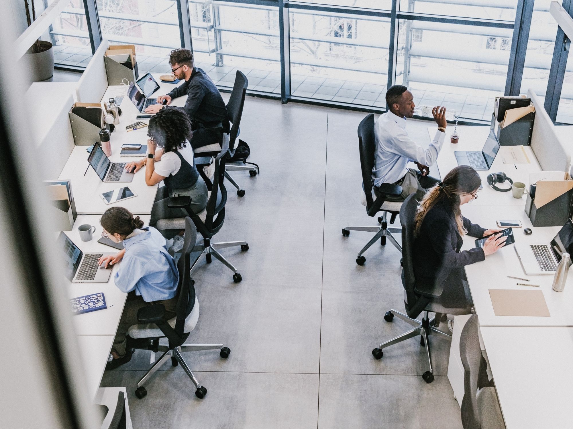 Several people are working in a modern open-plan office, seated at desks with laptops, notebooks, and office supplies, while some are focused on their computers and others are using tablets or taking a short break.