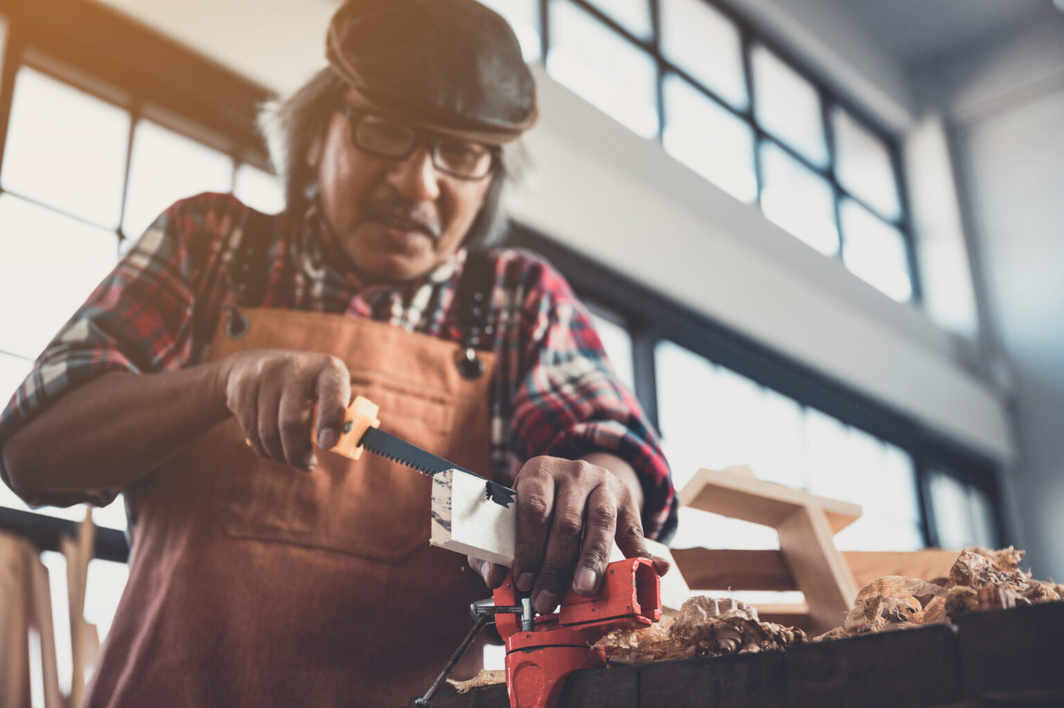 The image shows an older man wearing a cap, glasses, and a plaid shirt with an apron, working in a well-lit workshop. He is using a small hand saw to cut a piece of wood held securely in a red vise, with wood shavings and a small wooden structure visible on the workbench in front of him.