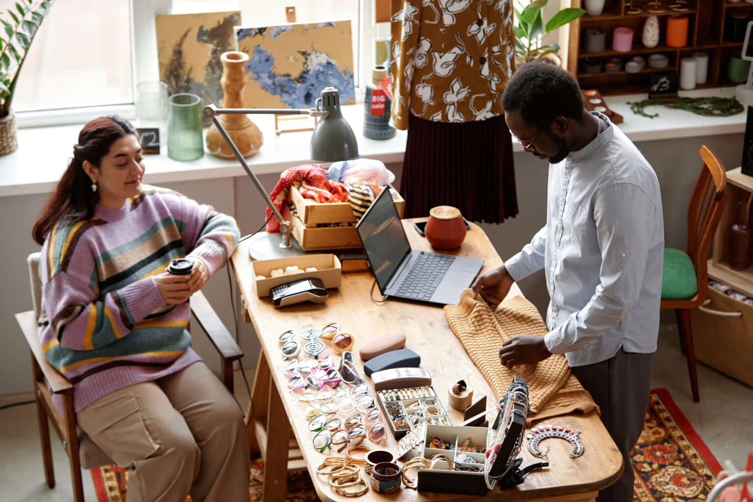A man and woman work together at a wooden table covered with jewelry, glasses, and accessories, while the man folds a textured fabric and the woman, wearing a striped sweater, sits holding a coffee cup.