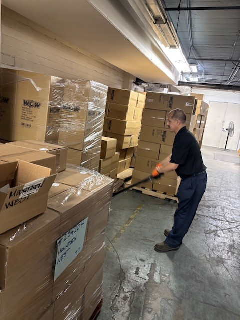 A worker uses a pallet jack to move a large stack of cardboard boxes in a warehouse aisle filled with tall piles of packaged goods.