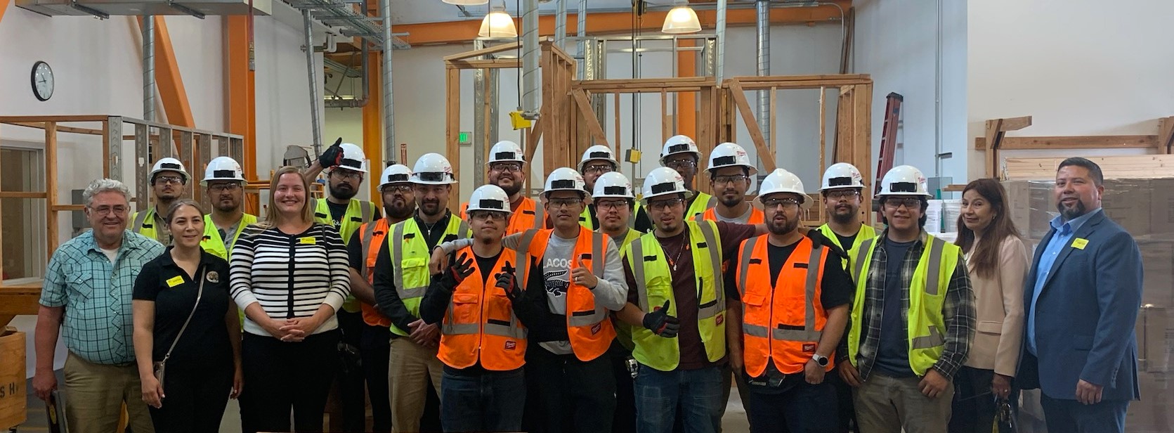 Group of people wearing hard hats and high-visibility safety vests posing together inside a construction training facility with wood framing structures in the background.