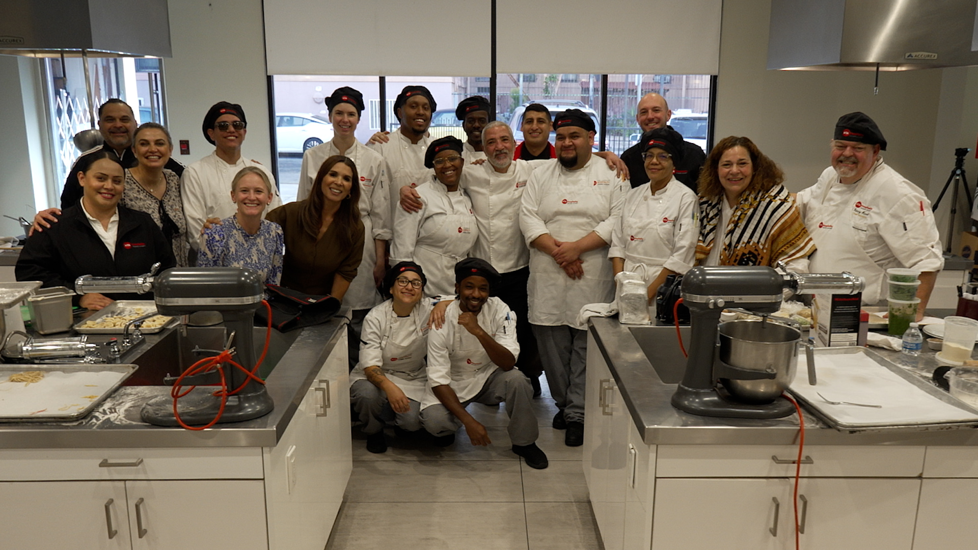 Group of people wearing chef coats and hats posing together in a culinary classroom kitchen with mixers and food preparation stations in the foreground.