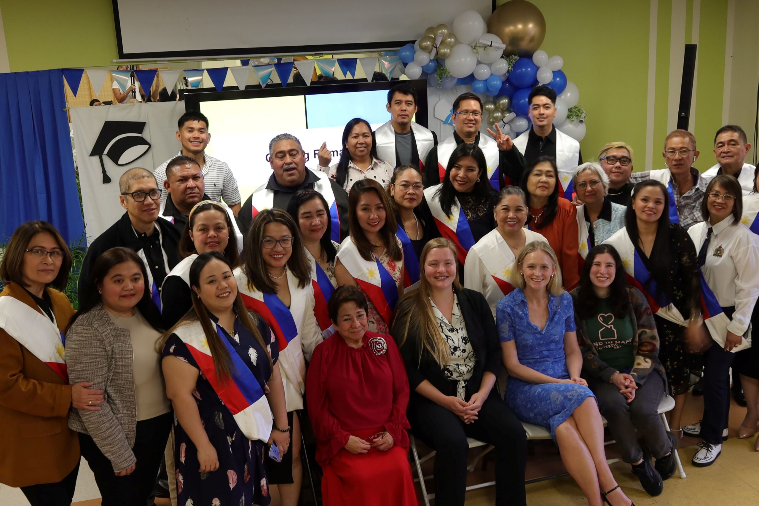 Large group of adults posing indoors for a group photo, many wearing sashes in red, blue, and white, with balloons and a presentation screen in the background.